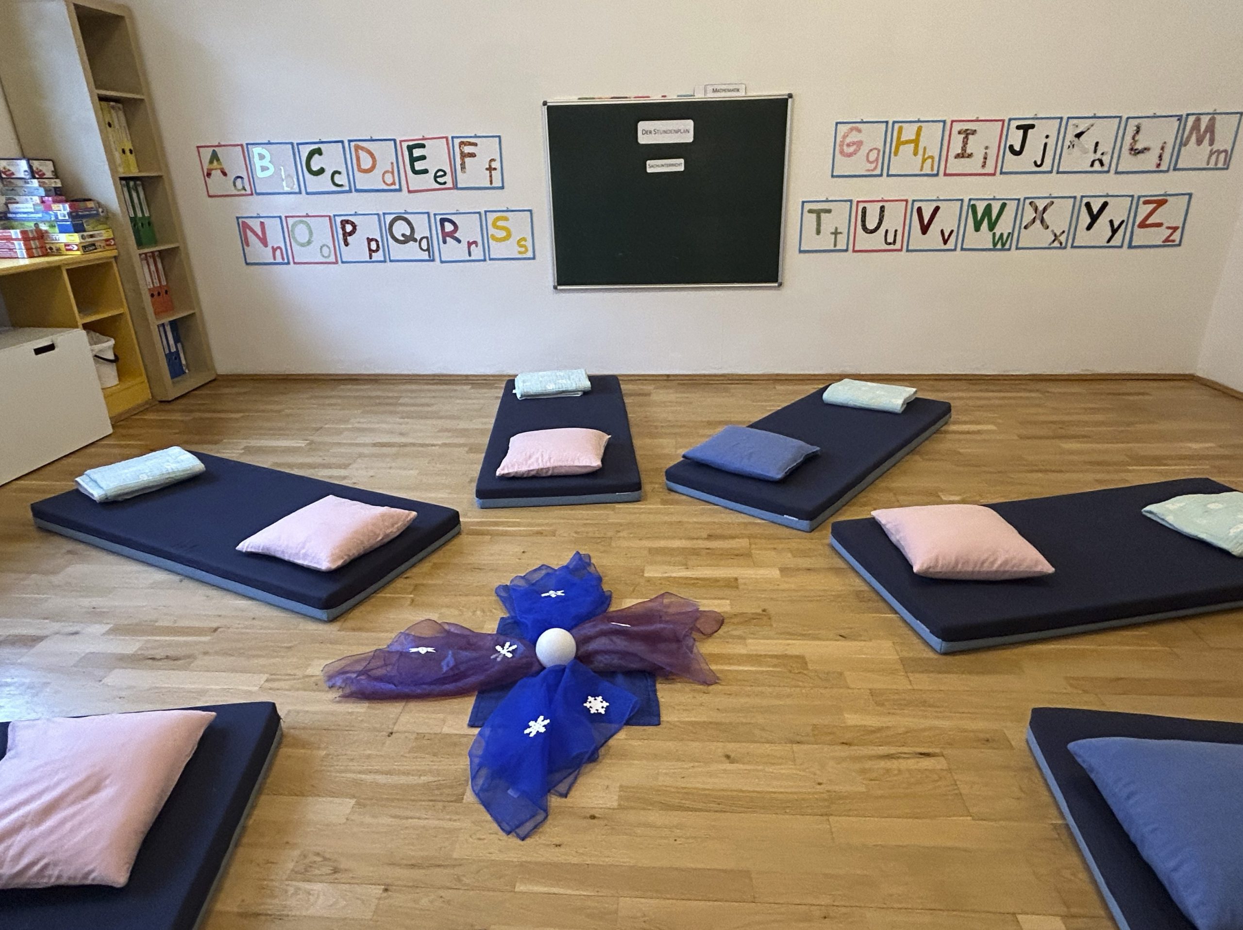 Tafel an der Wand, ABC in Buchstaben an Wand, Matratzen mit Pölstern und Decken liegen in einem Kreis am Boden