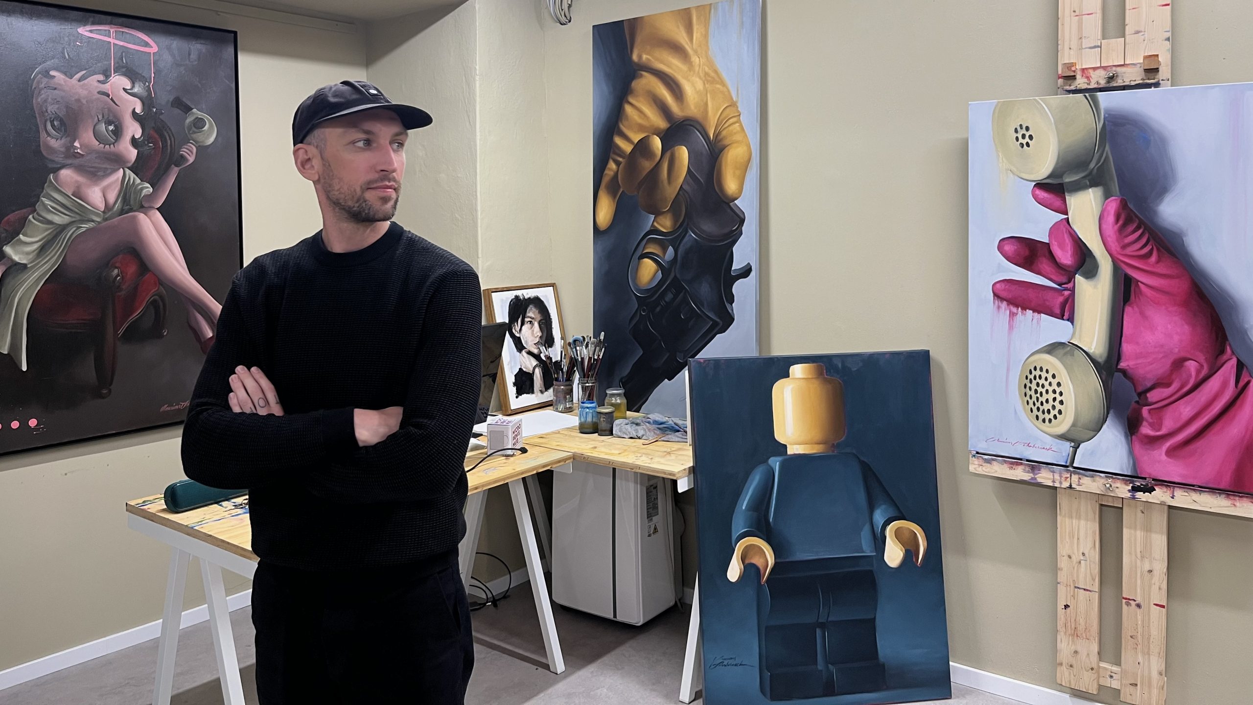 Es ist der Künstler Monsieur Schabernack mit einigen seiner Werke im Hintergrund zu sehen. Er trägt eine schwarze Kappe und schaut nach rechts.