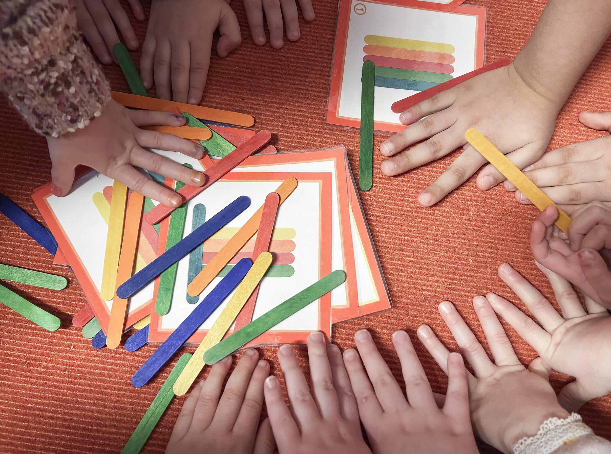 Zuordnungsspiel mit bunten Holzstäbchen und Bildern in der Mitte eines orangenen Teppichs umgeben von Kinderhänden die in die Mitte zum Spiel greifen.