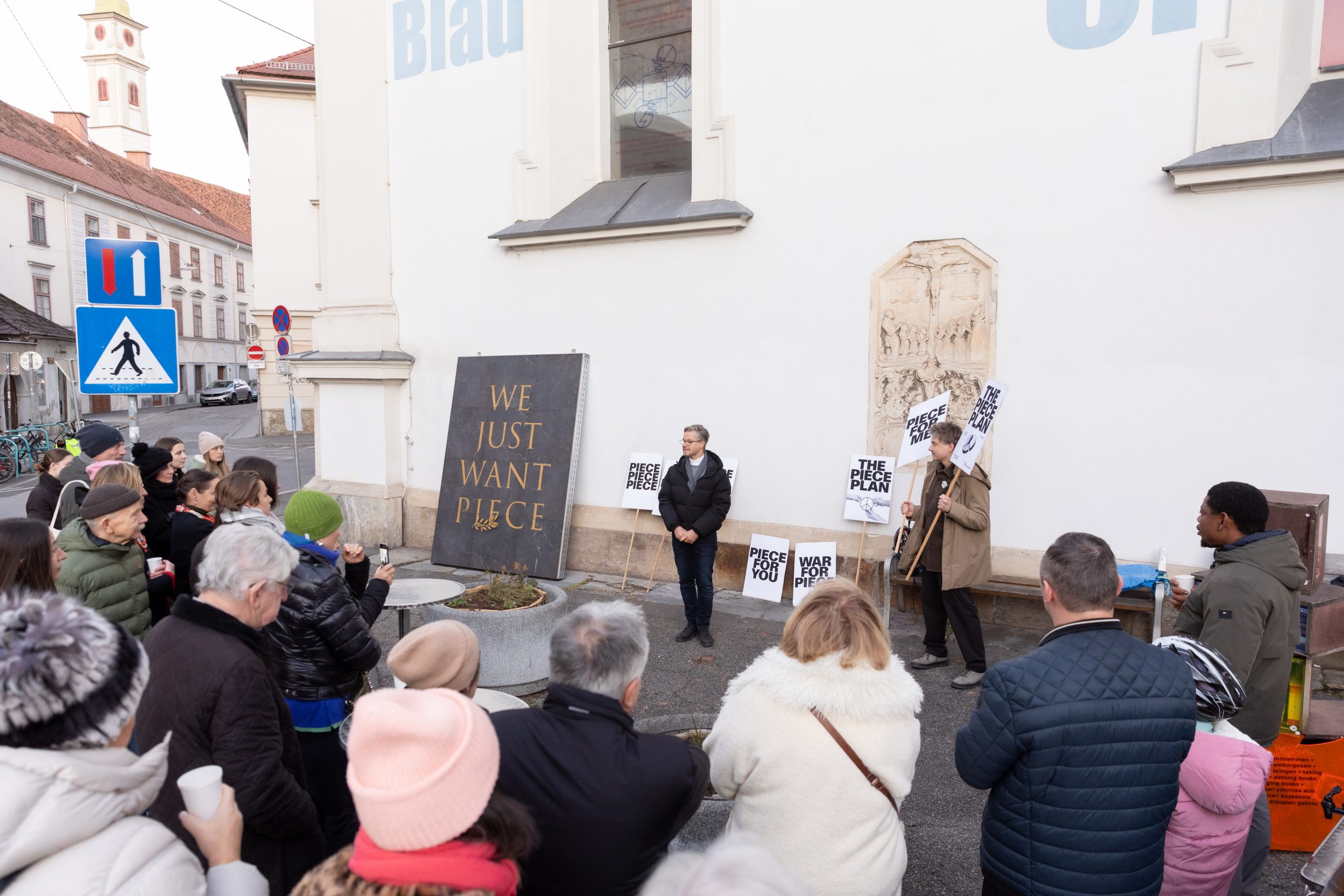 Eine Menschenmasse vor einer Kirche betrachten eine große Tafel mit der Aufschrift „We just want piece". Daneben stehen zwei Männer mit kleinen Schildern, auf denen kurze Phrasen mit Aufforderungen für Frieden stehen.