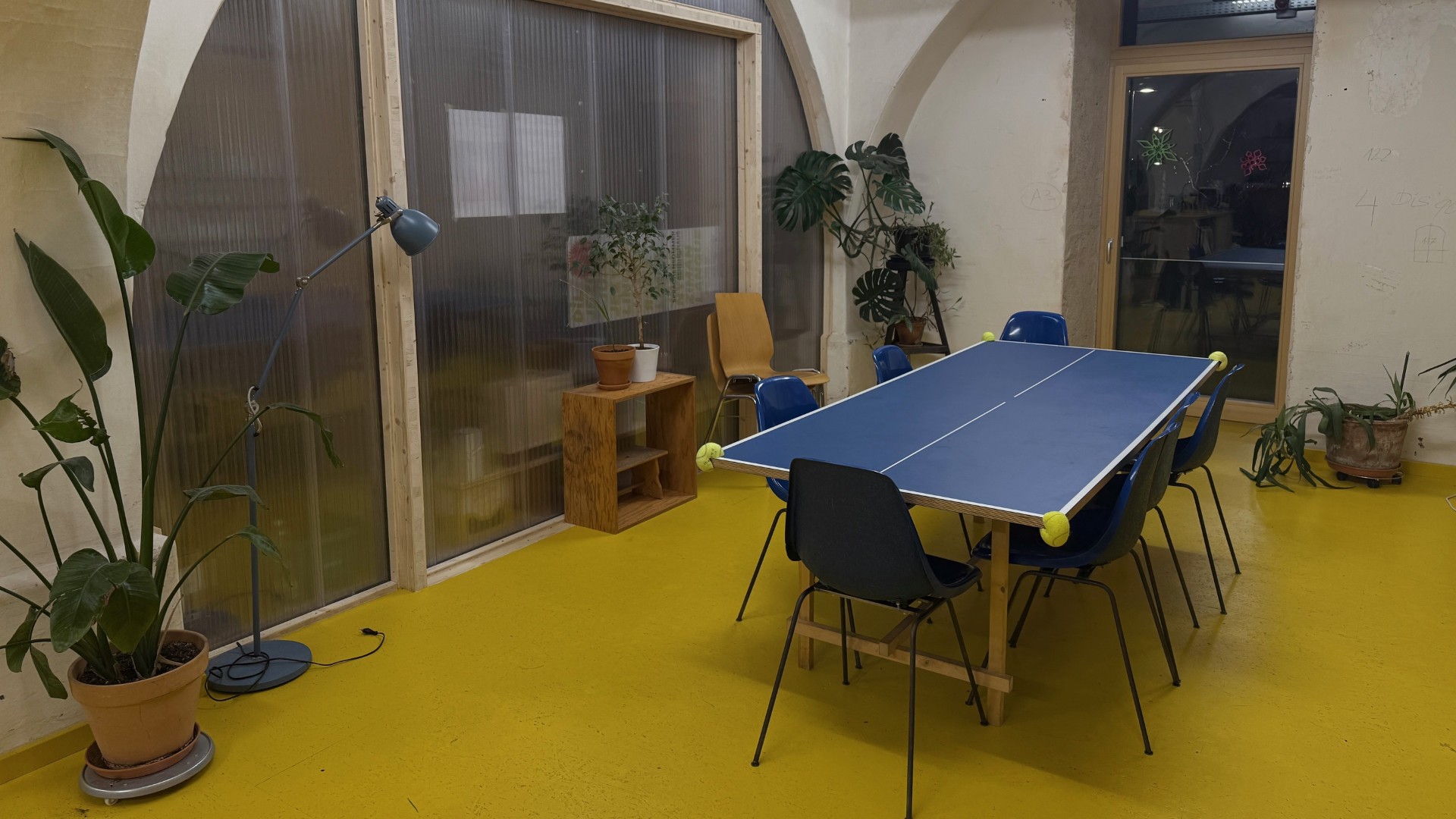 Ein Raum mit einem Tisch in der Mitte mit sechs blauen Stühlen. Gelber Boden, mehrere Pflanzen stehen in den Ecken. Der Tisch war einst ein Ping-Pong-Tisch, aber das Netz in der Mitte wurde entfernt und die Tischbeine ausgetauscht.