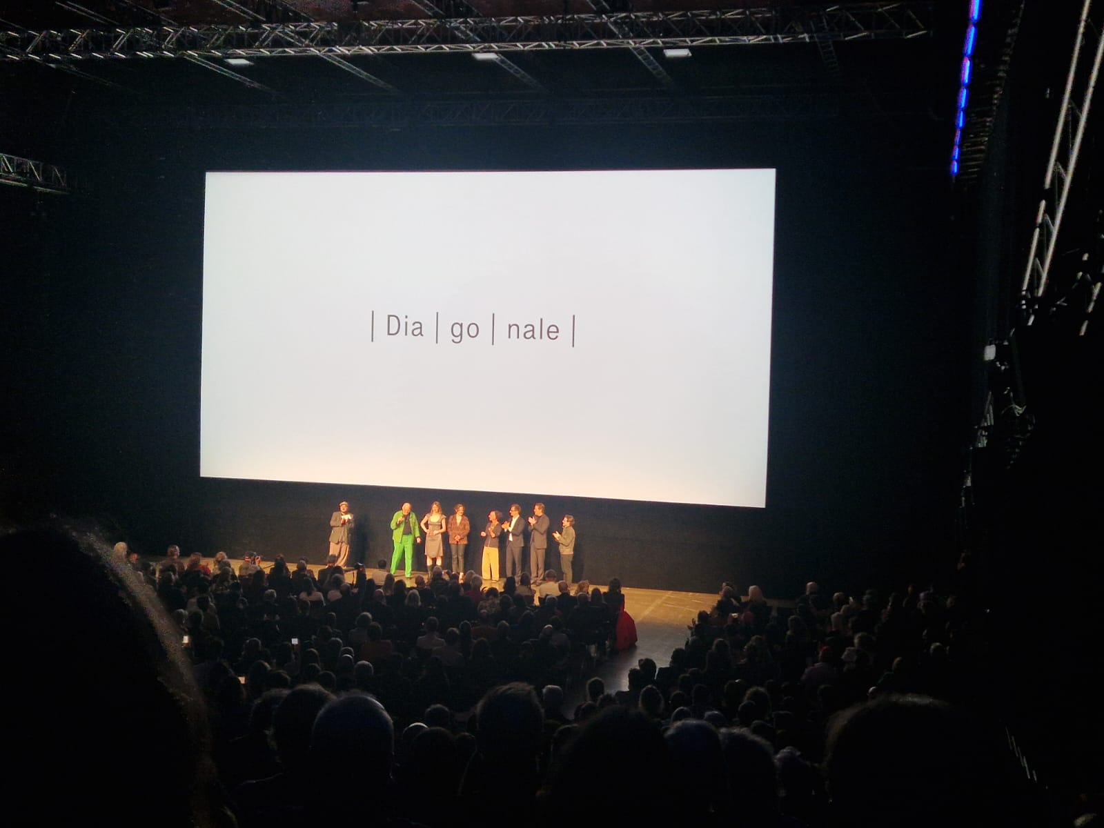 Auf dem Bild ist eine große Leinwand mit der Schrift „Diagonale" drauf und davor steht die Filmcrew des Eröffnungsfims „Rose".