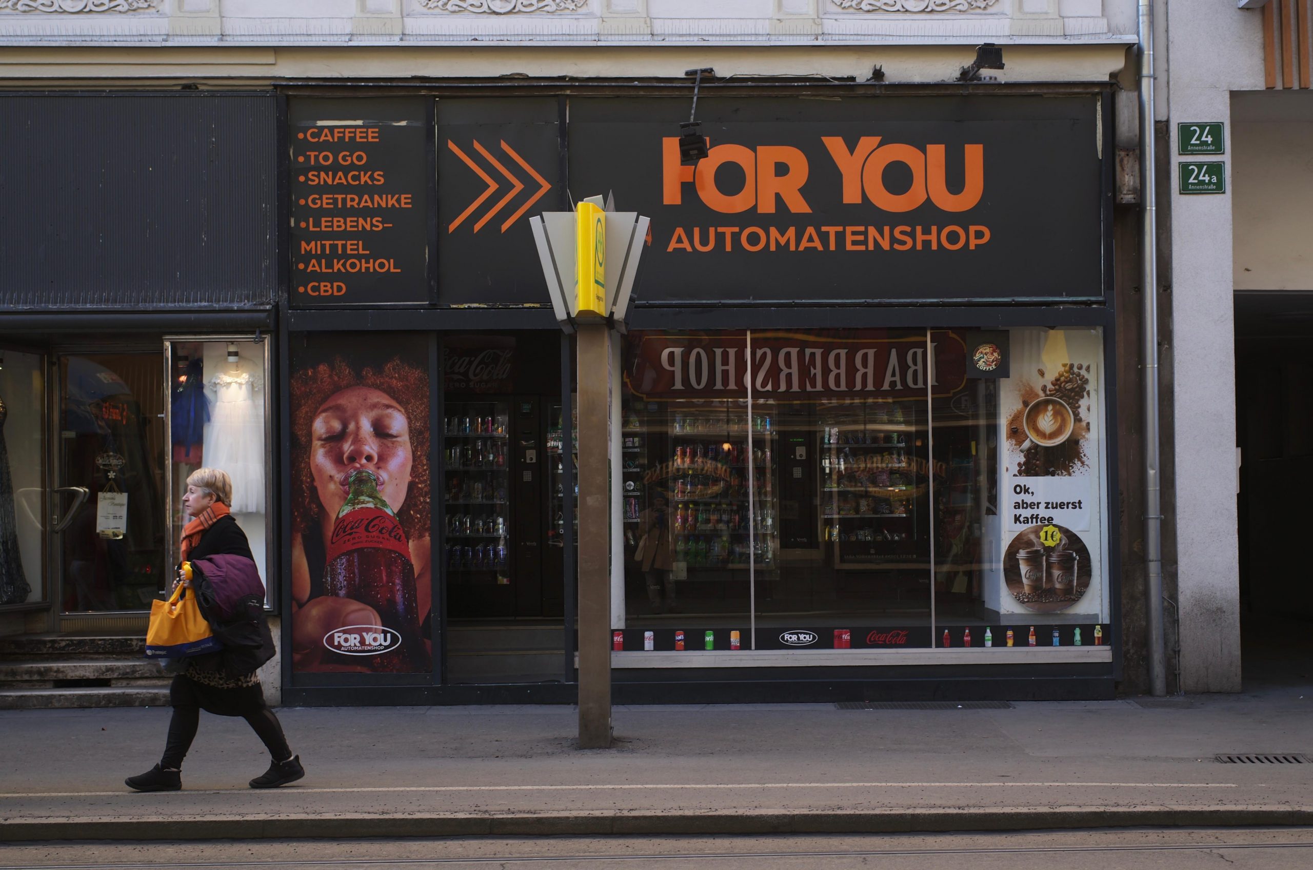 Eine Schaufensterfront eines Automatenshops, die obere Hälfte der Front ist schwarz mit oranger Schrift. Vor allem der Name des Shops „For You Automatenshop" ist besonders groß. Davor ist ein Haltestellenschild und eine Frau, die gerade daran vorbeigeht.