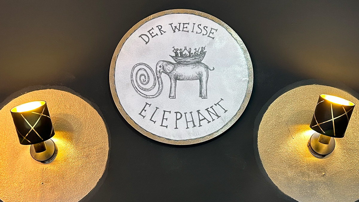 Auf einer schwarzen Wand, ist in der Mitte das weiße Logo mit schwarzer Schrift zu sehen, das einen Elefanten zeit, der ein Boot mit Musikern am Rücken trägt. Links und rechts davon zwei goldene Kreise mit schwarzen Wandlampen, die das goldene Licht unterstreichen