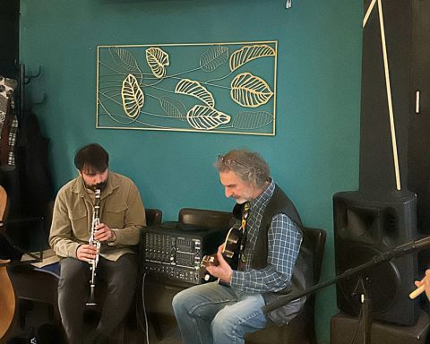 Vier Musiker aus unterschiedlichen Ländern musizieren mit ihren Instrumenten. Ganz links der Musiker mit Kontrabass, daneben sitzend der Klarinettenspieler, dazwischen steht ein Verstärker, als nächstes sitzt der Gitarrenspieler mit Gitarre und ganz rechts vor dem Mikrofon der Musiker mit der traditionellen Flöte "Ney"