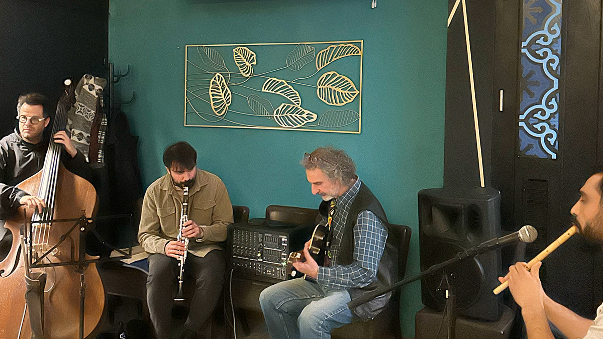 Vier Musiker aus unterschiedlichen Ländern musizieren mit ihren Instrumenten. Ganz links der Musiker mit Kontrabass, daneben sitzend der Klarinettenspieler, dazwischen steht ein Verstärker, als nächstes sitzt der Gitarrenspieler mit Gitarre und ganz rechts vor dem Mikrofon der Musiker mit der traditionellen Flöte "Ney"