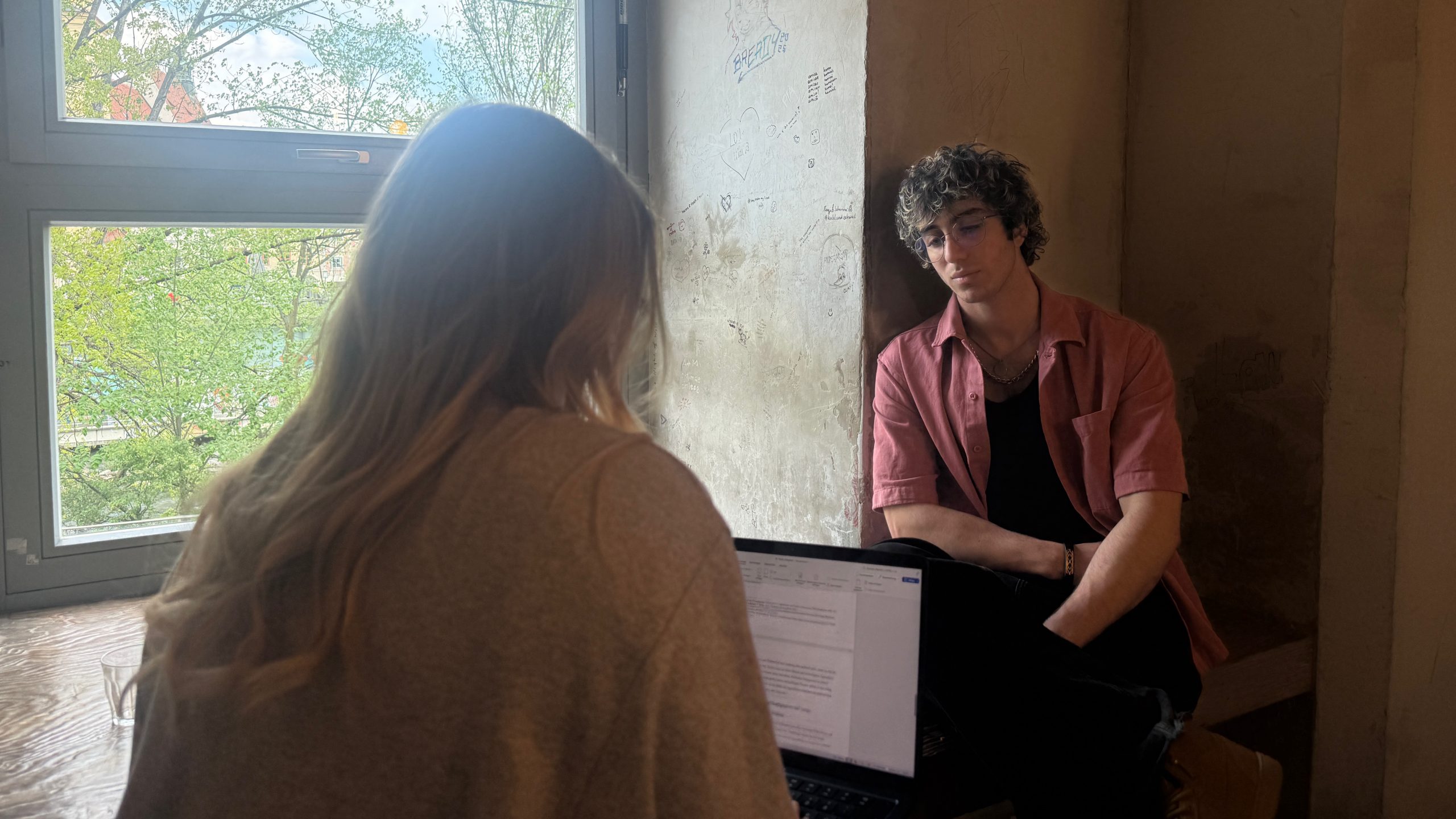 Samiko sitzt neben einem Fenster, der Blick gesenkt. Ihm gegenüber sieht man die Interviewerin der Annenpost von hinten mit einem Laptop am Schoß.