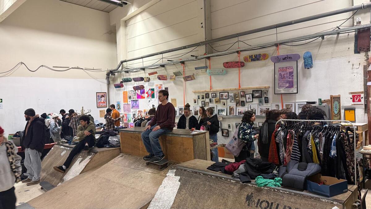 Viele Menschen stehen in der Skatehalle vor Kleiderstangen und Skate-Hindernissen, die als Ablage für Kleidungsstücke verwendet werden. An der weißen Wand hängen mehrere bunte Skateboards und viele Poster und gerahmte Bilder.