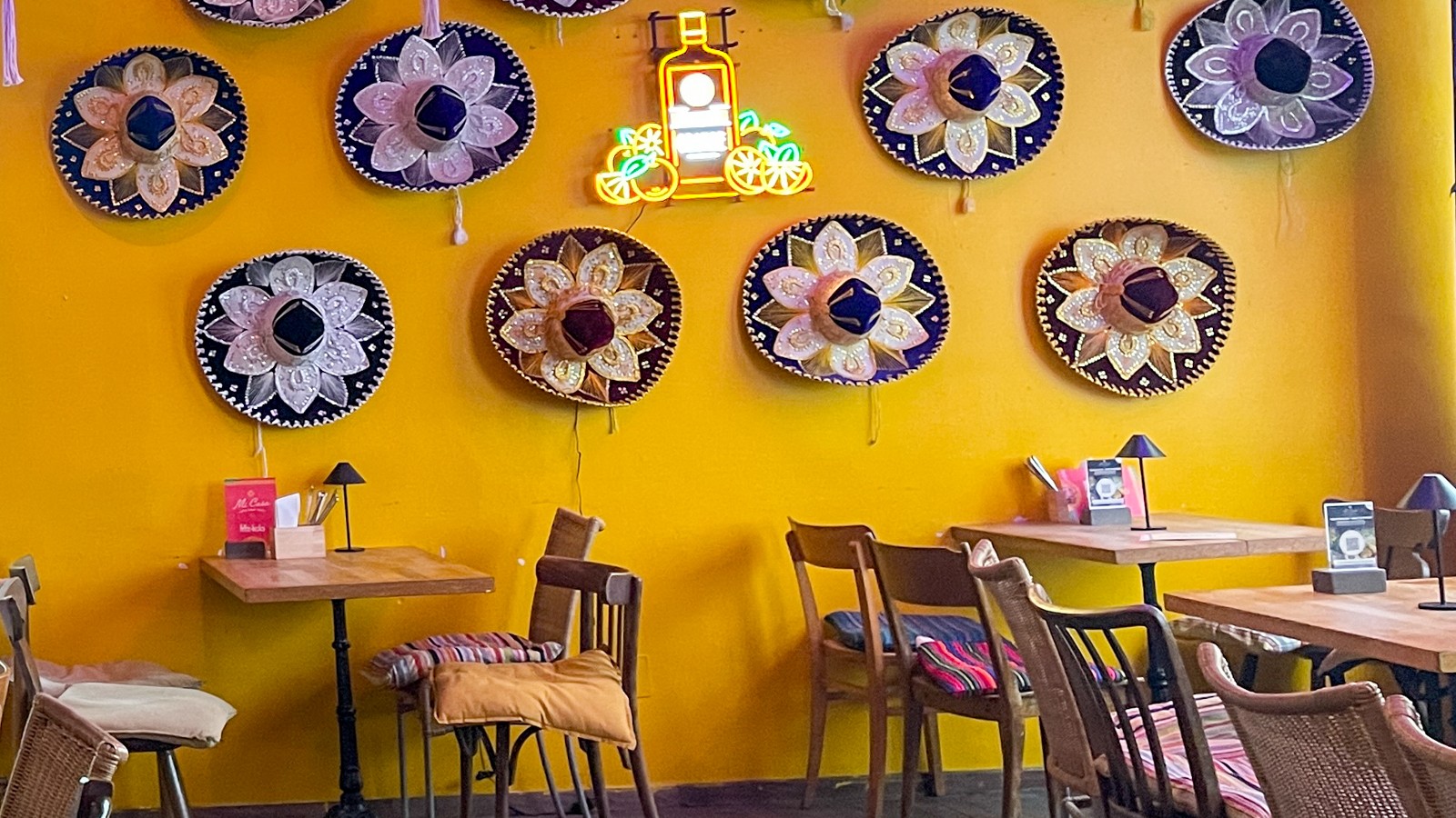 Die Sitzplätze im Mi Casa mit hölzernen Stühlen und Tischen, hinten eine gelbe Wand dekoriert mit Sombreros