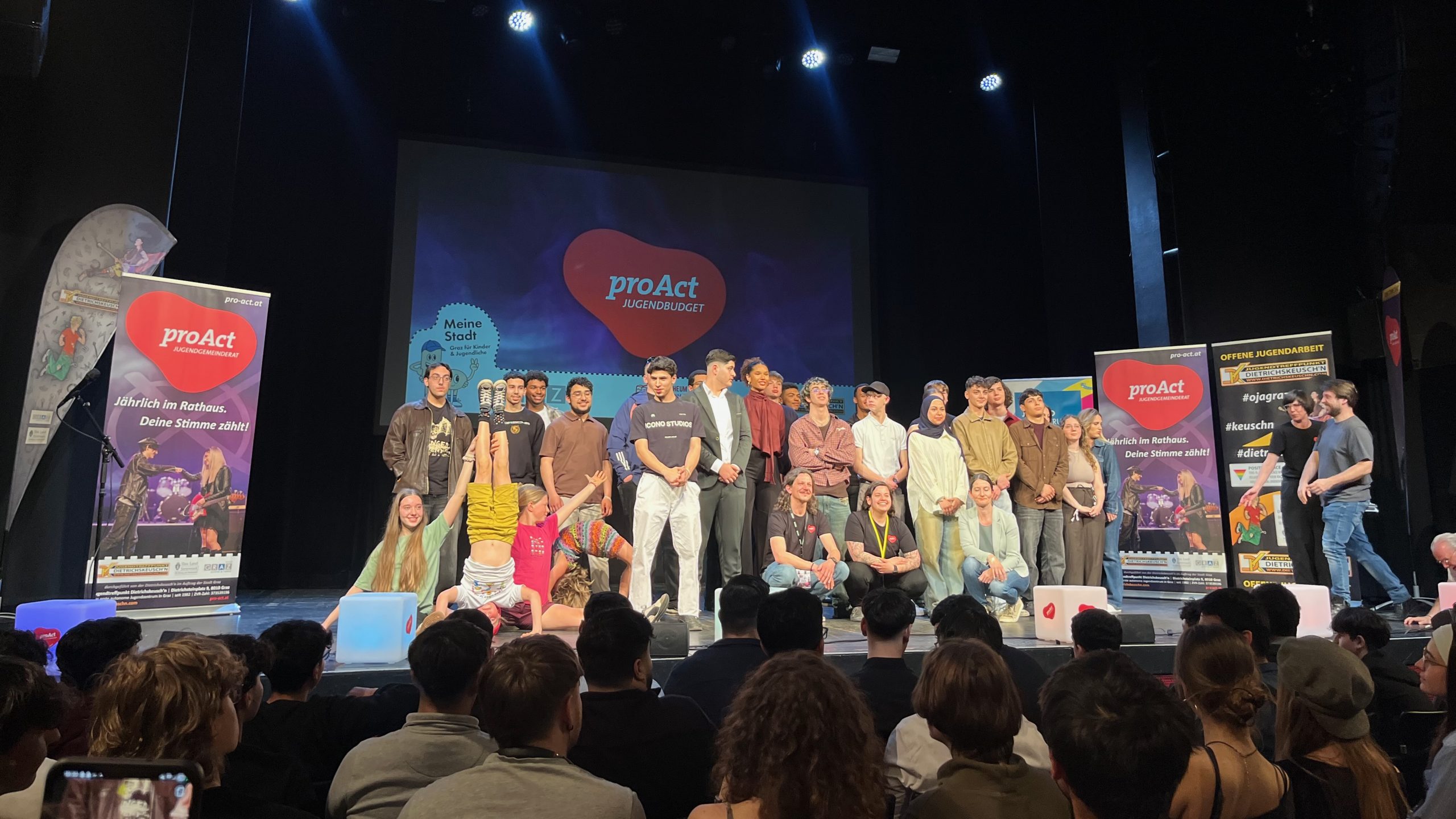 Eine Gruppe Jugendlicher steht auf einer Bühne, im Hintergrund und auf den Seiten sind Poster mit dem Schriftzug „proAct" zu sehen.