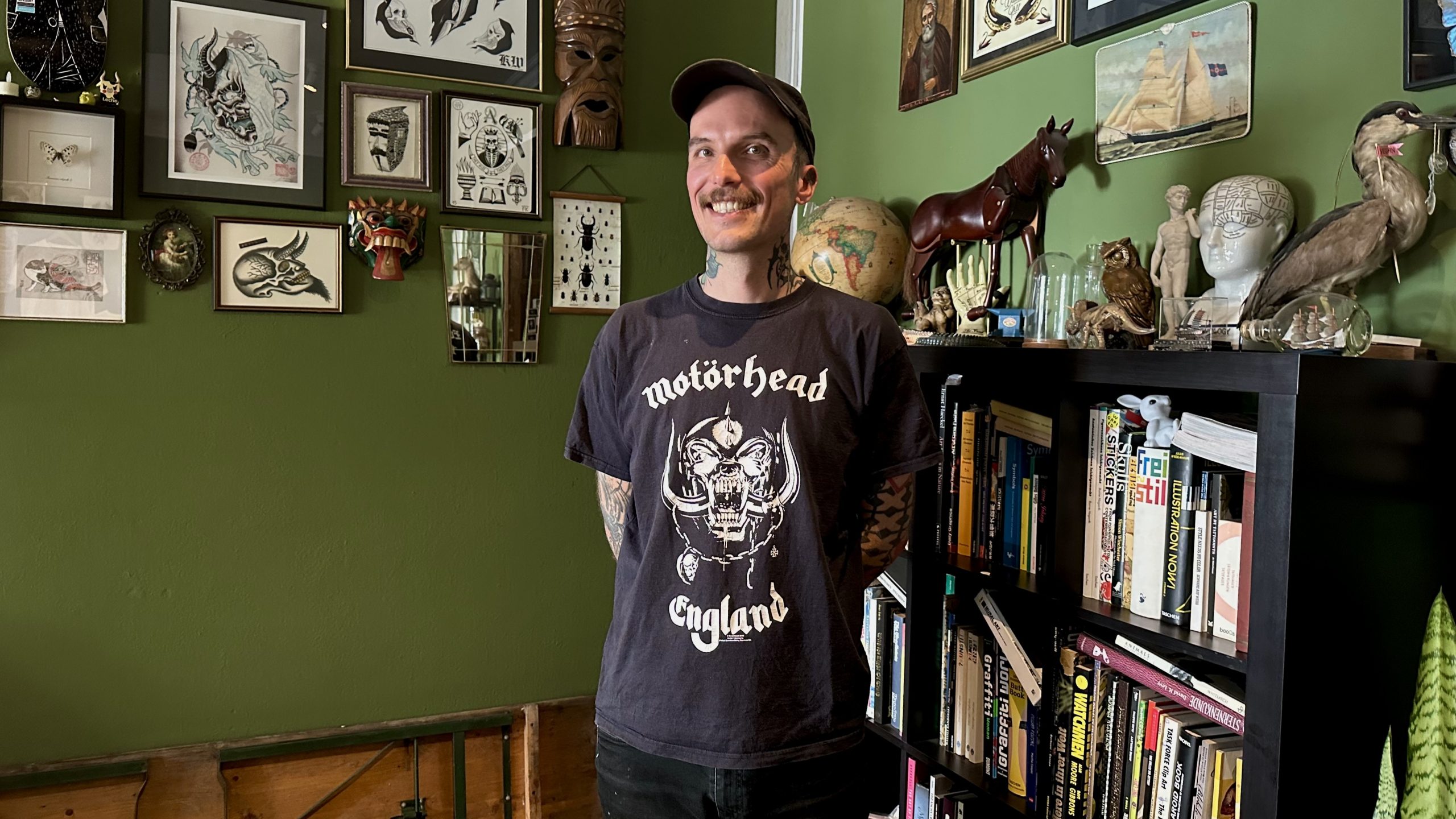 Der Tätowierer Adrian Mair in seinem Studio „Three Nails Tattoo“. Er trägt eine Cap und ein schwarzes Outfit. Viele grafische Bilder hängen an der grünen Wand. Das Regal ist voll mit Büchern.