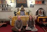 Ein älterer Mann und eine junge Frau sitzen in einem farbenfrohen Raum vor einem buddhistischen Altar.