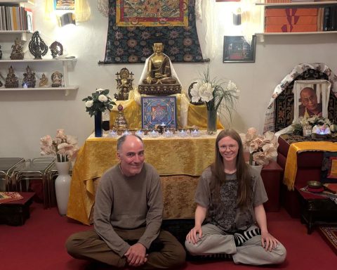 Ein älterer Mann und eine junge Frau sitzen in einem farbenfrohen Raum vor einem buddhistischen Altar.